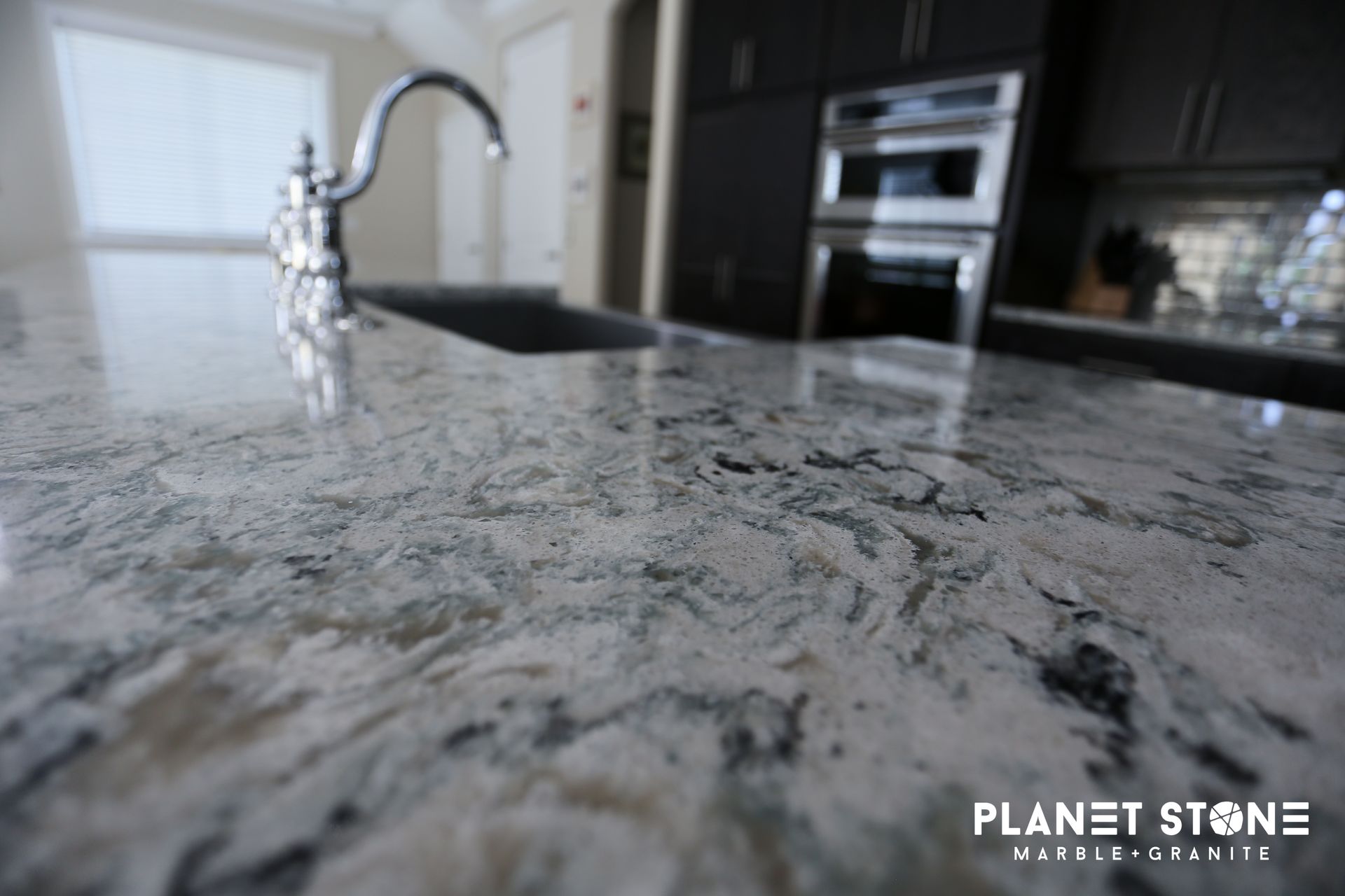 Close-up of a polished gray granite kitchen countertop with sink and built-in oven in the background