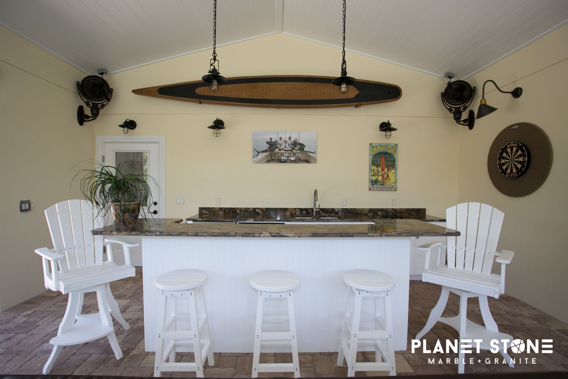 Bright outdoor kitchen bar with white stools and chairs, surfboard light fixture, and dartboard on beige wall