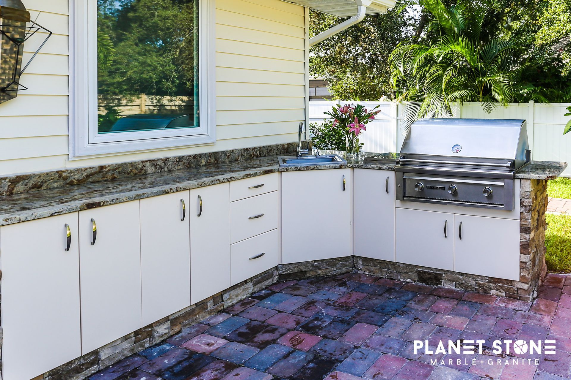 Outdoor L-shaped stone kitchen with white cabinets, granite countertop, sink, and grill beside a house