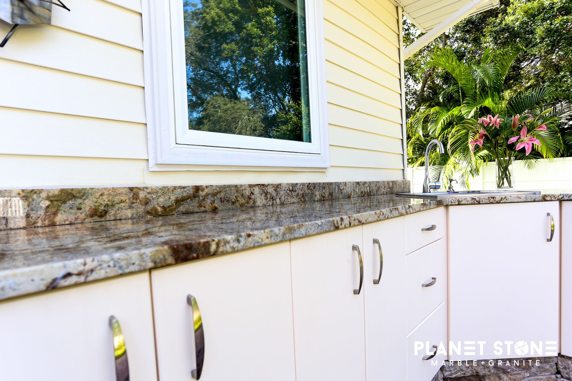 White outdoor cabinets with stone countertop beneath a window on a house exterior.