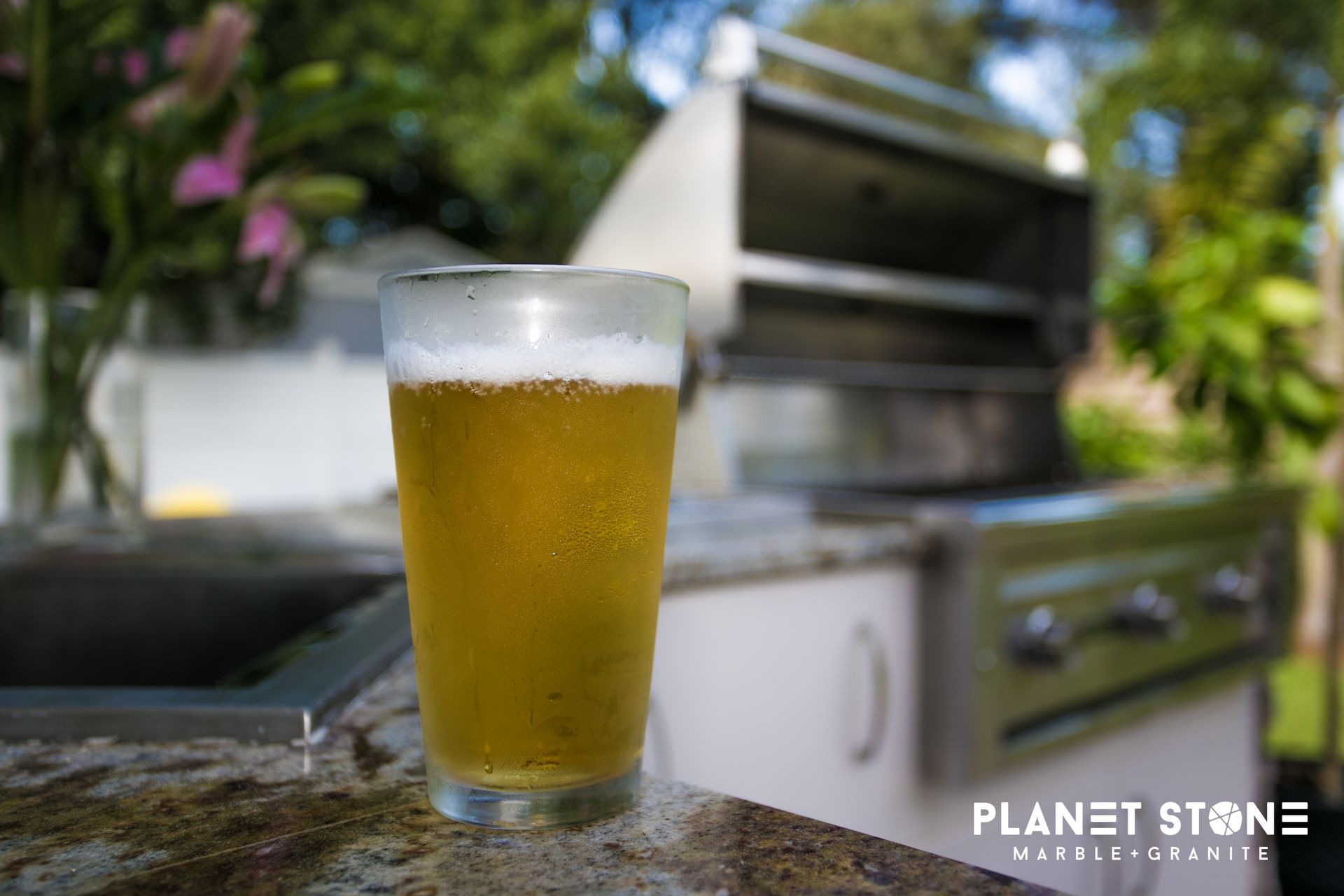 A frosty beer glass on a patio table beside an outdoor grill in a sunny backyard.