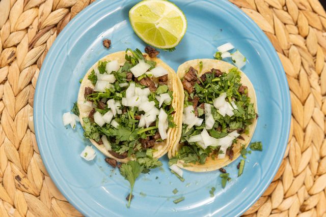 Two tacos with cilantro and diced onion on a blue plate, with a lime wedge on a woven placemat.