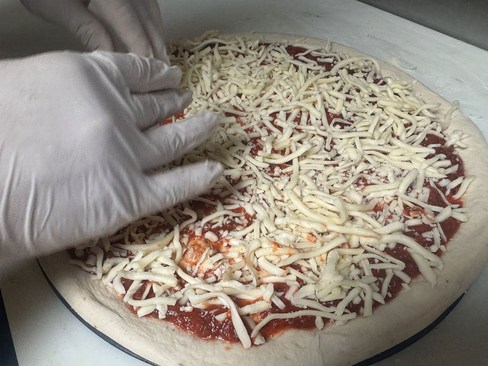 Hands wearing gloves spreading shredded cheese on a pizza.