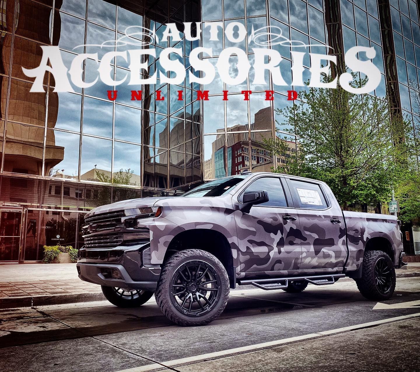 Camouflage truck with black wheels parked in front of a modern building; 