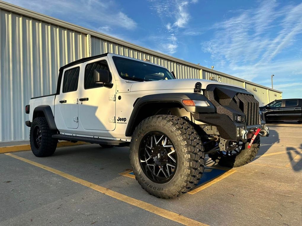 White Jeep Gladiator truck with black wheels and a custom front bumper parked in front of a metal building.
