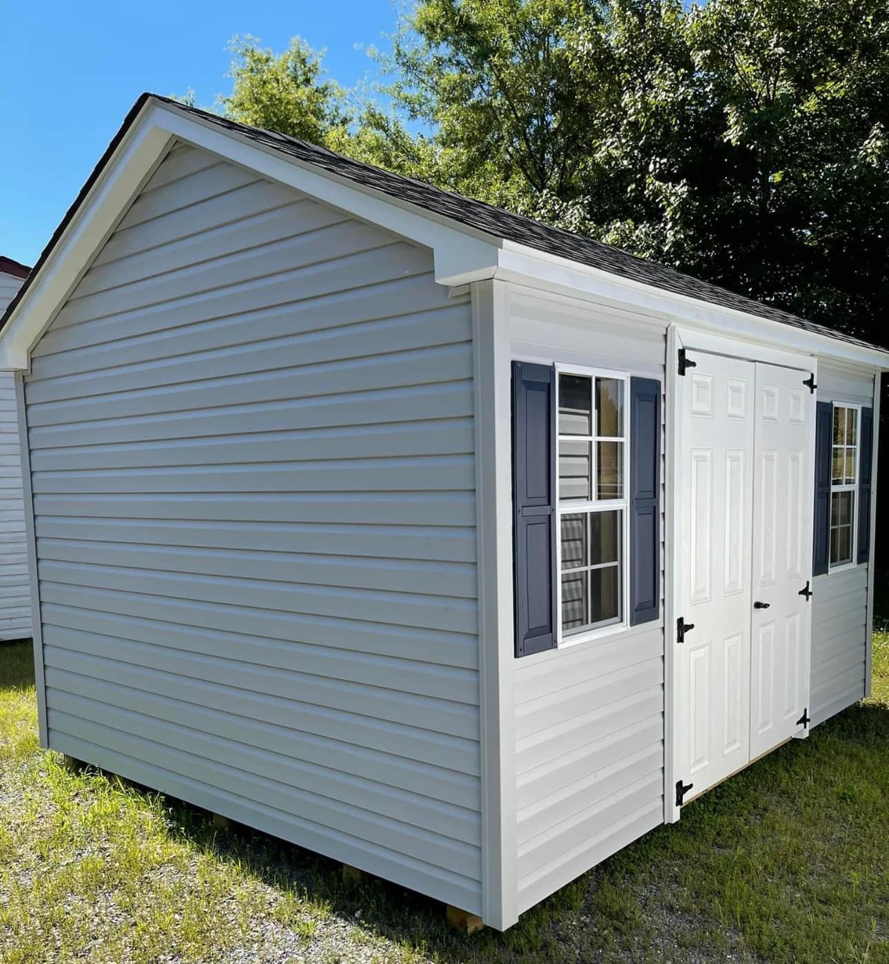 A white shed with black shutters is sitting in the grass.
