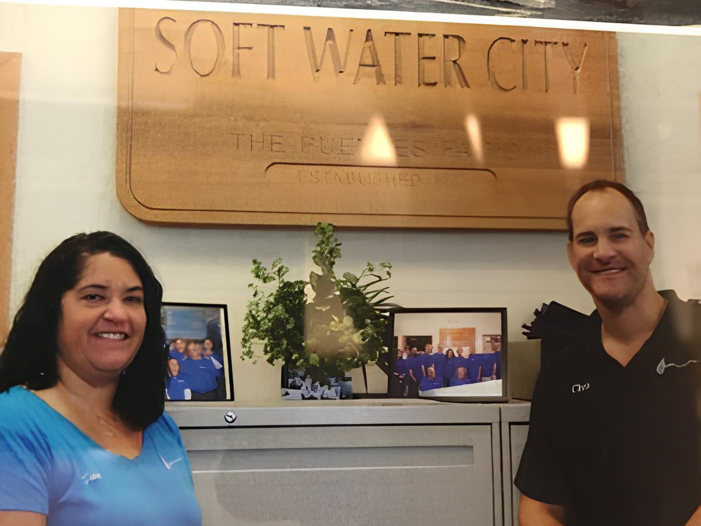 Two smiling people stand in front of a Soft Water City sign, with photos and a plant displayed on a cabinet.