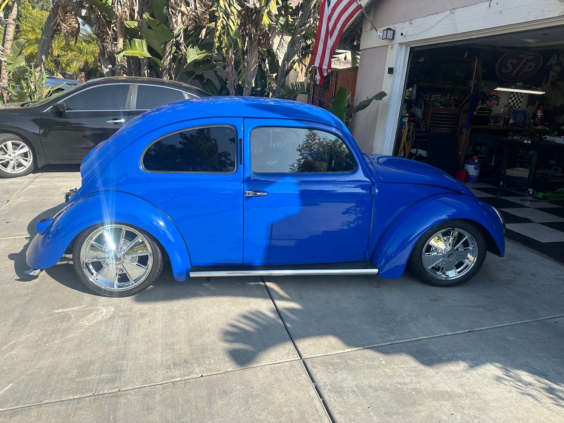 Blue Volkswagen Beetle with chrome wheels parked in front of a garage, a black car beside it.