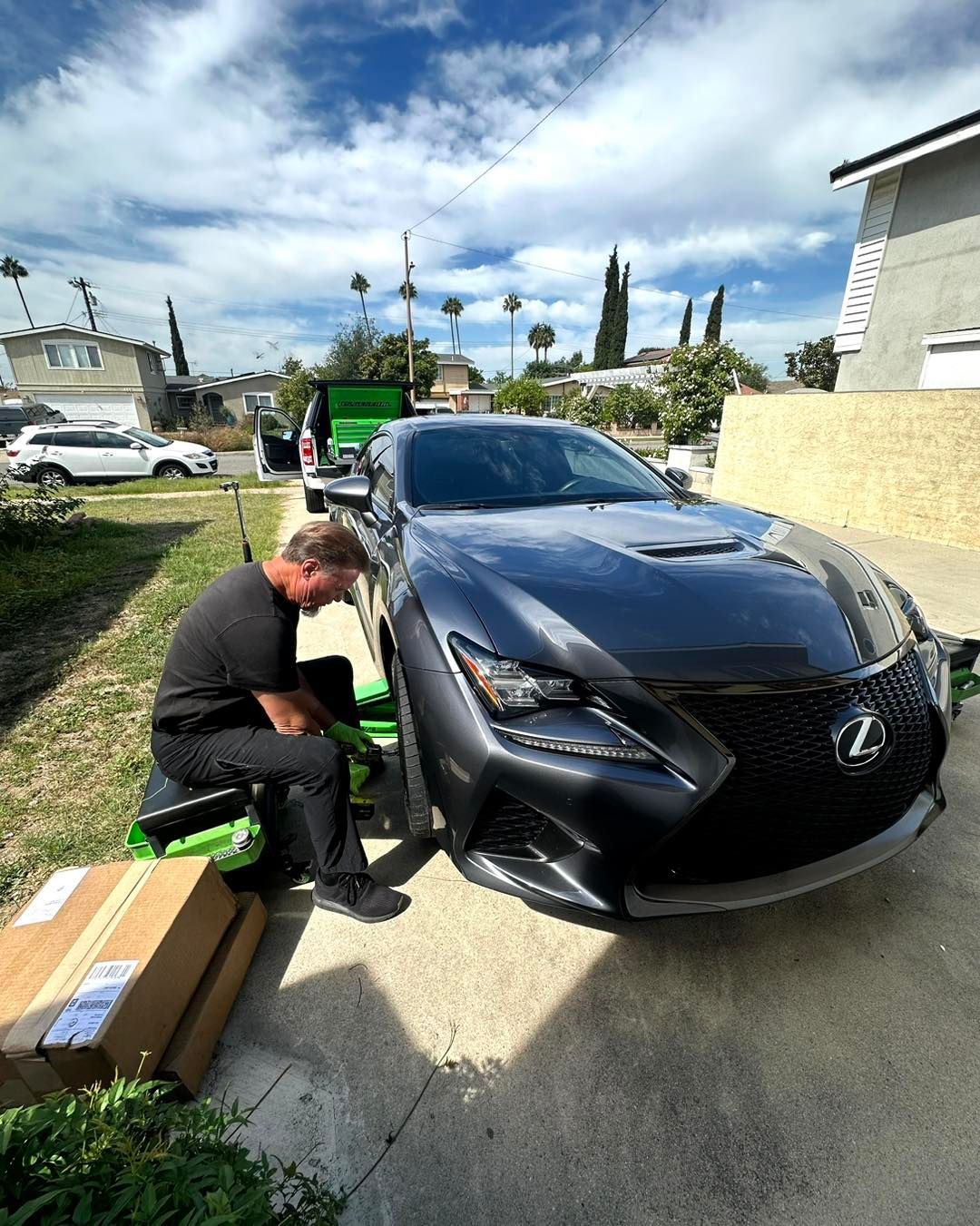 Man working on a dark gray Lexus in a driveway on a sunny day. Boxes and tools are nearby.
