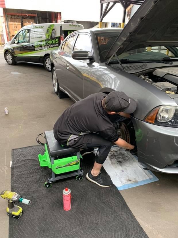 A mechanic working on the front wheel of a gray car with the hood open, sitting on a green stool, outside a shop.
