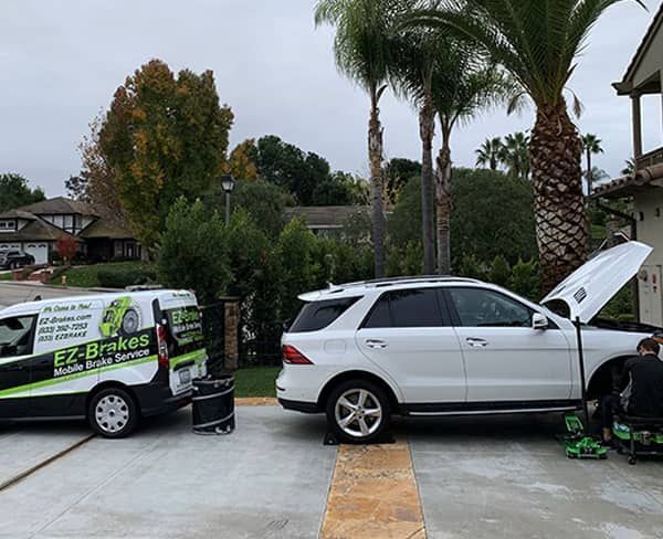 A white Mercedes SUV with open hood being serviced by a mobile brake service van in a driveway.