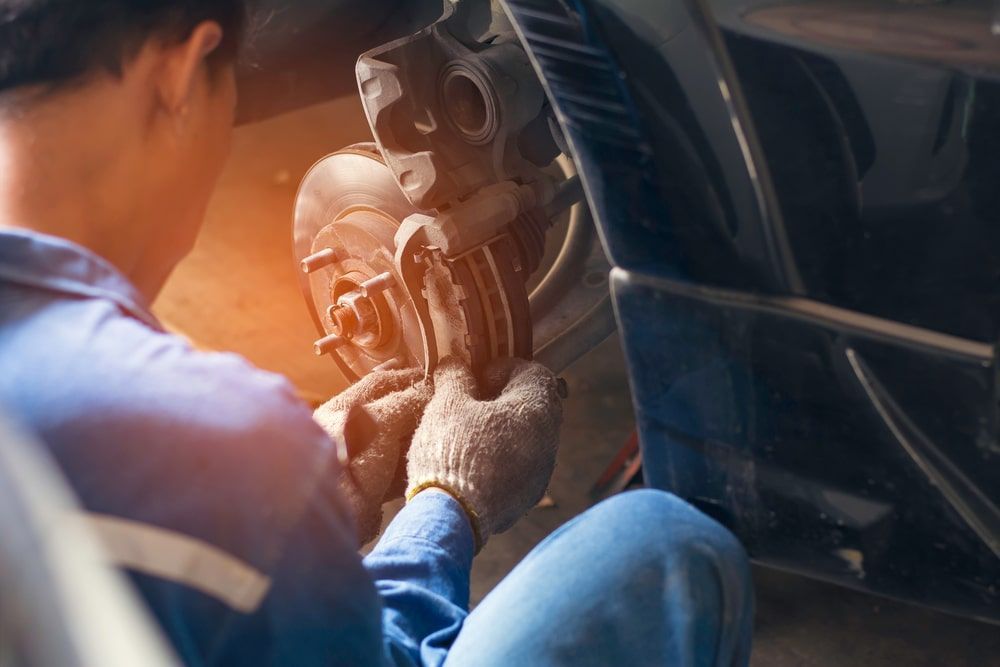 Mechanic working on car's brake system, wearing gloves and blue jumpsuit, in a garage setting.