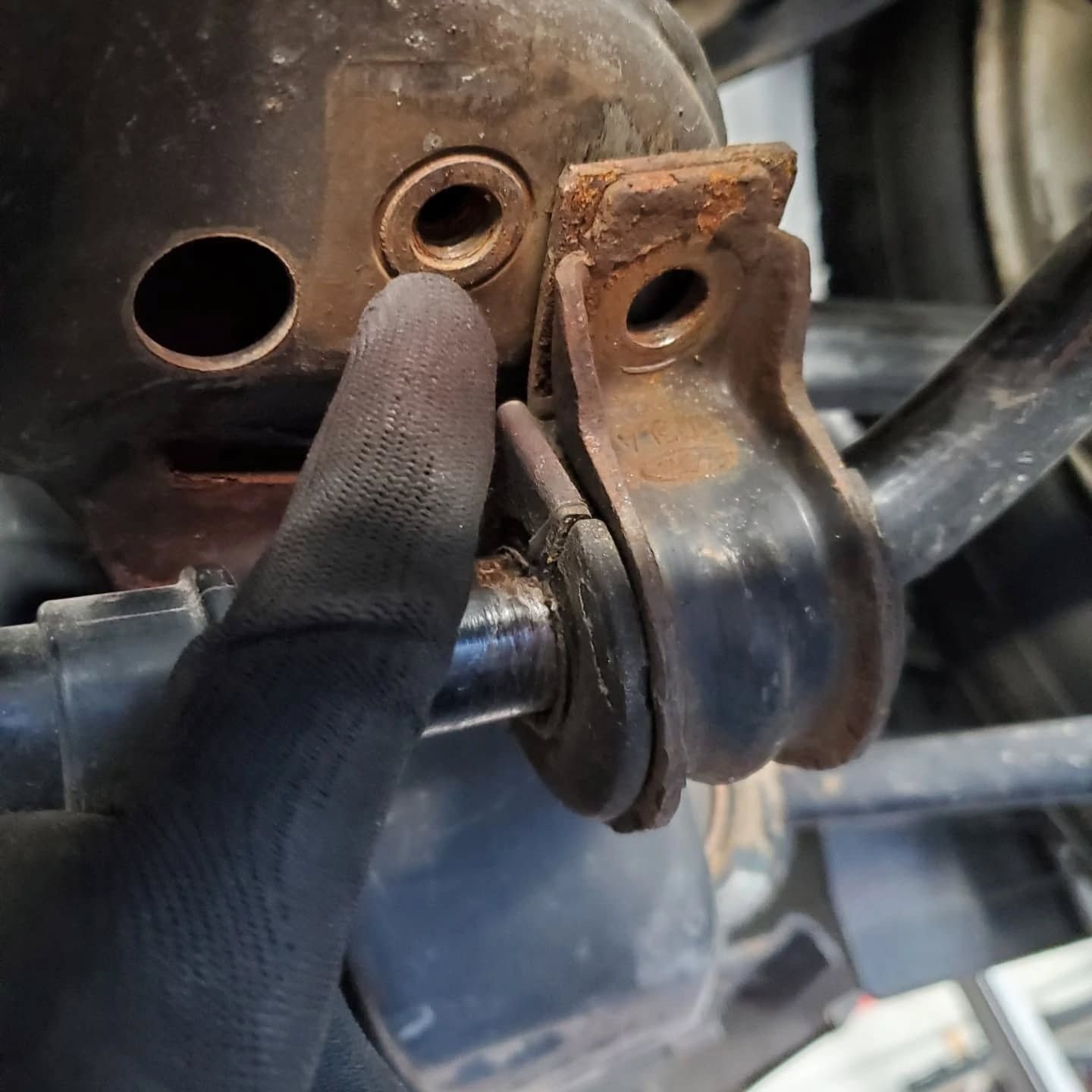 Gloved finger points to a rusty bolt hole on a metal vehicle suspension bracket.