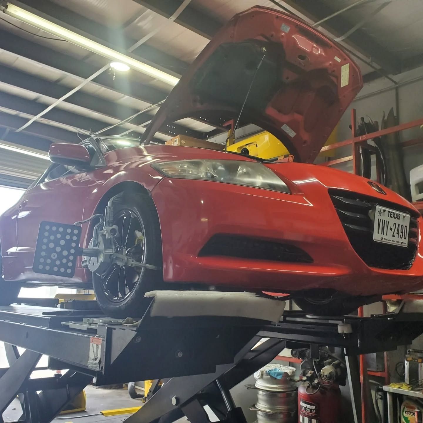 Red car on a car lift in a garage, hood open with exposed front end.