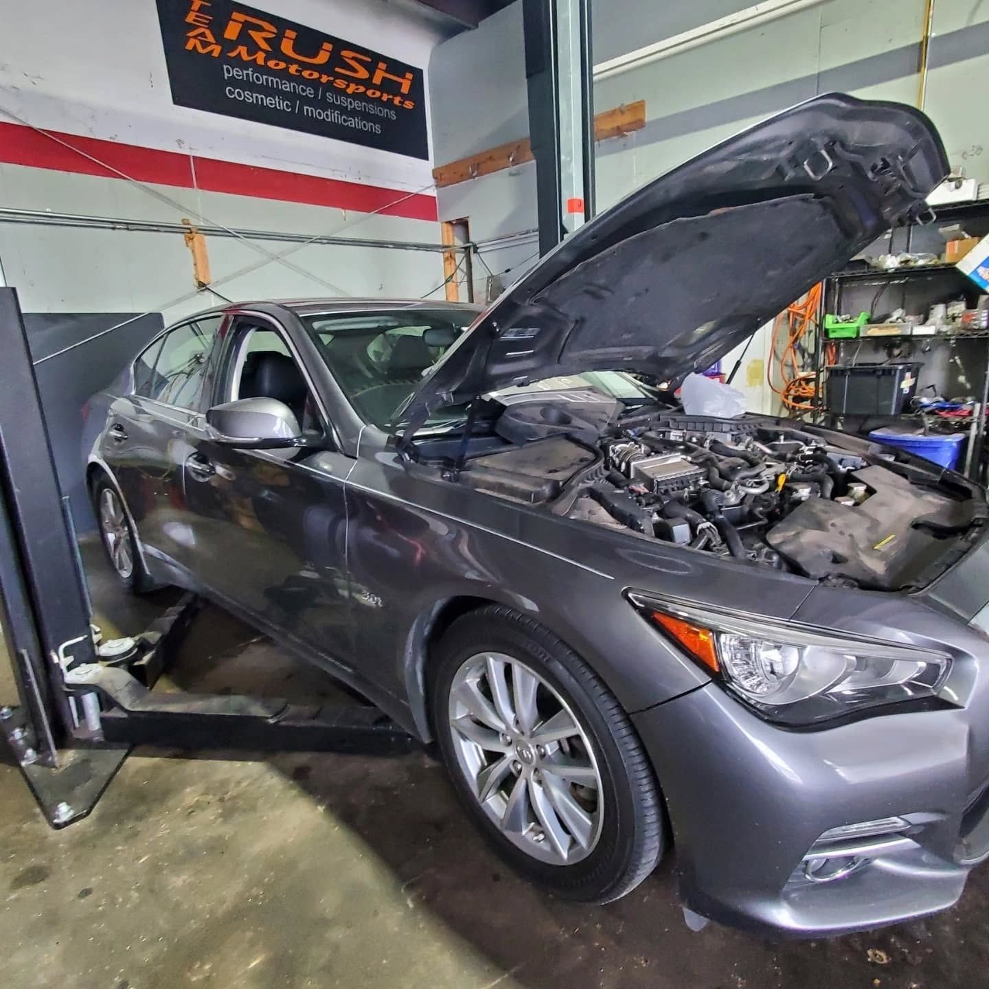 Gray sedan with hood open in an auto repair shop, showing engine bay and lift equipment.
