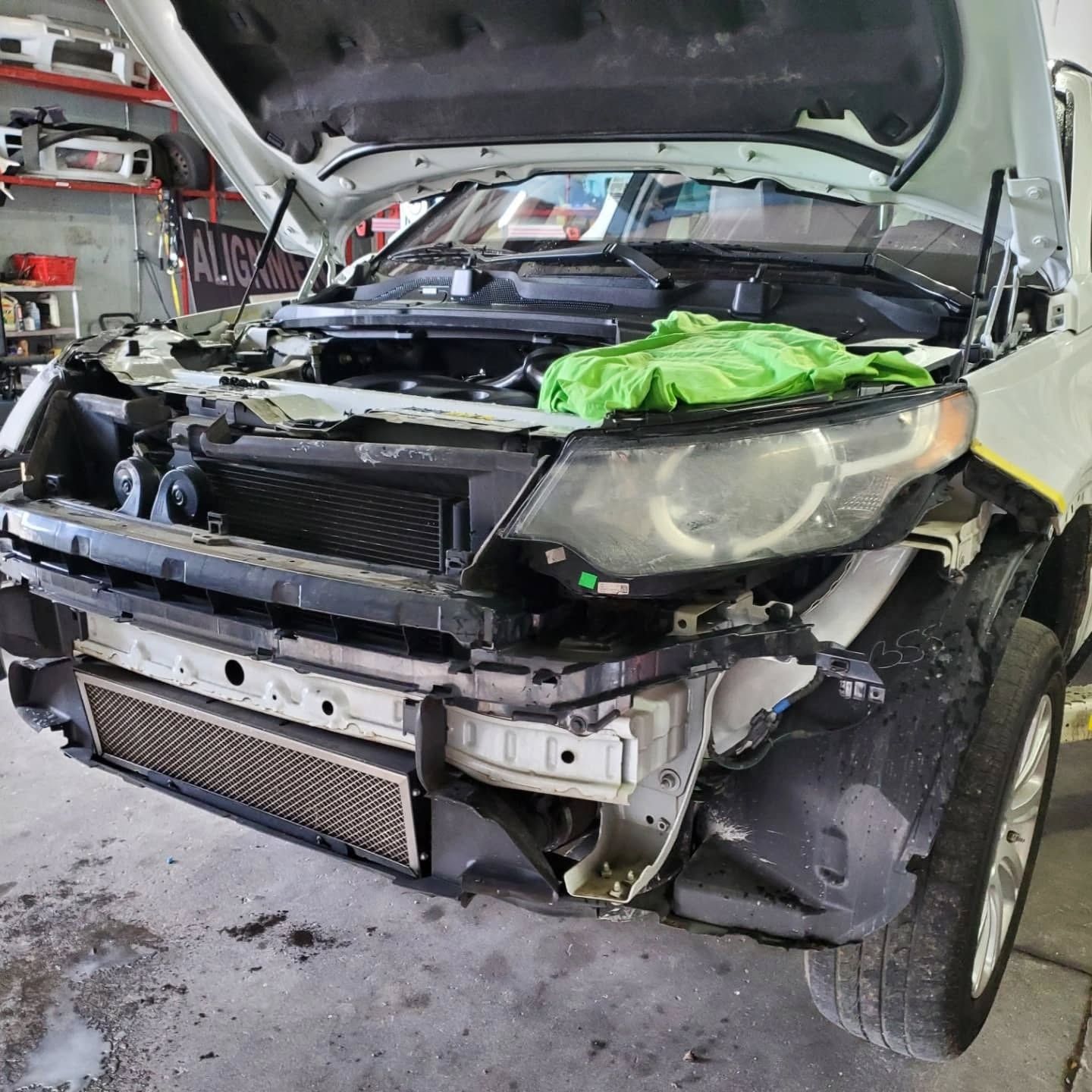 Severely damaged black car in a repair shop, hood open and front bumper removed.