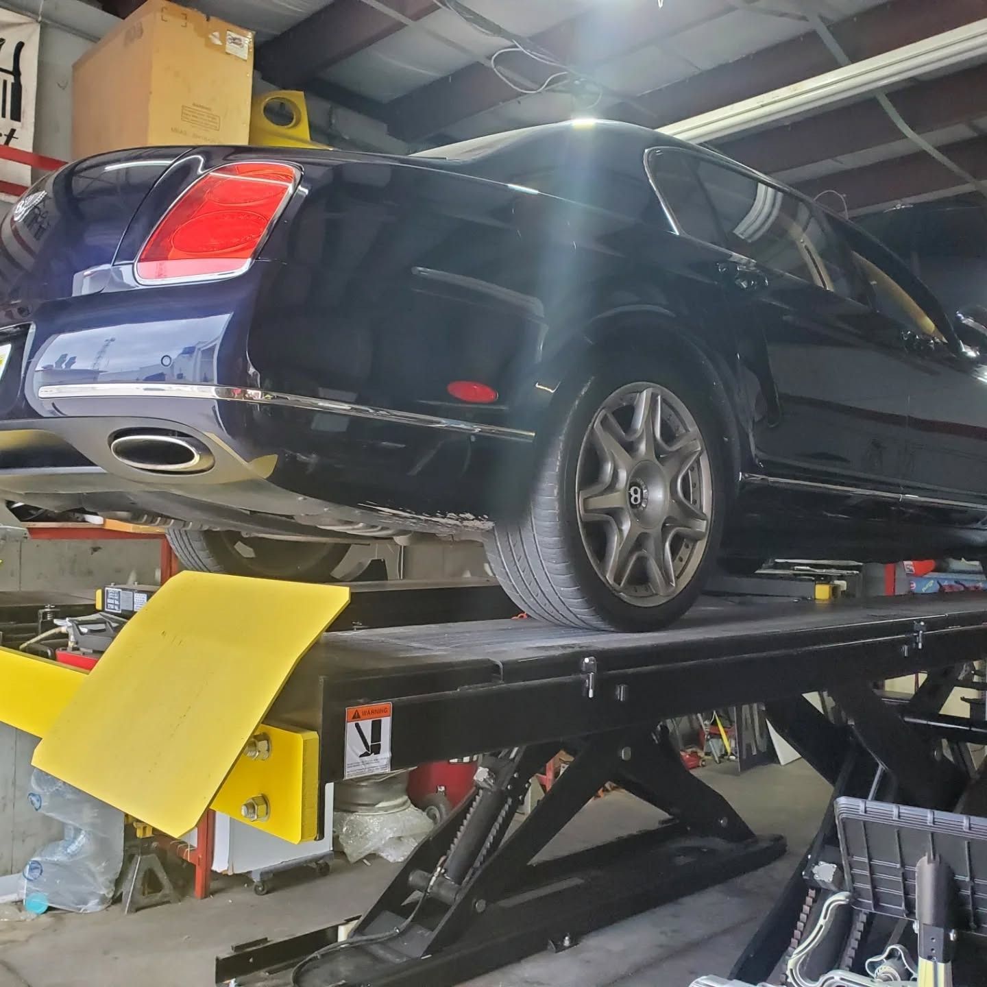 Black sedan on a garage lift in a repair shop, rear end raised with yellow support pad visible