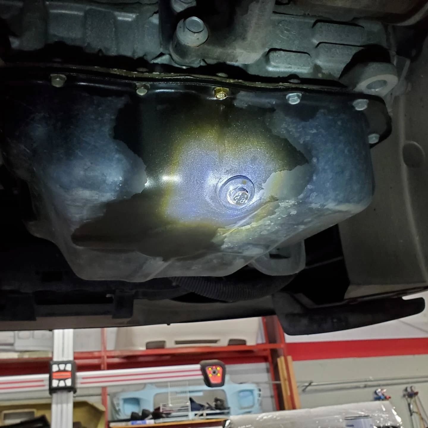 Undercarriage view of a car’s oil pan with a drain plug, showing oil residue in a garage lift bay