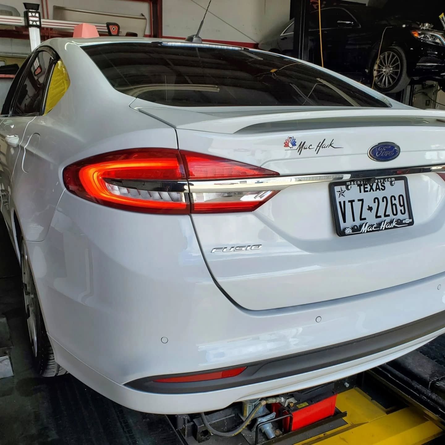 White Ford sedan on a shop lift, rear view in a garage with a Texas license plate.