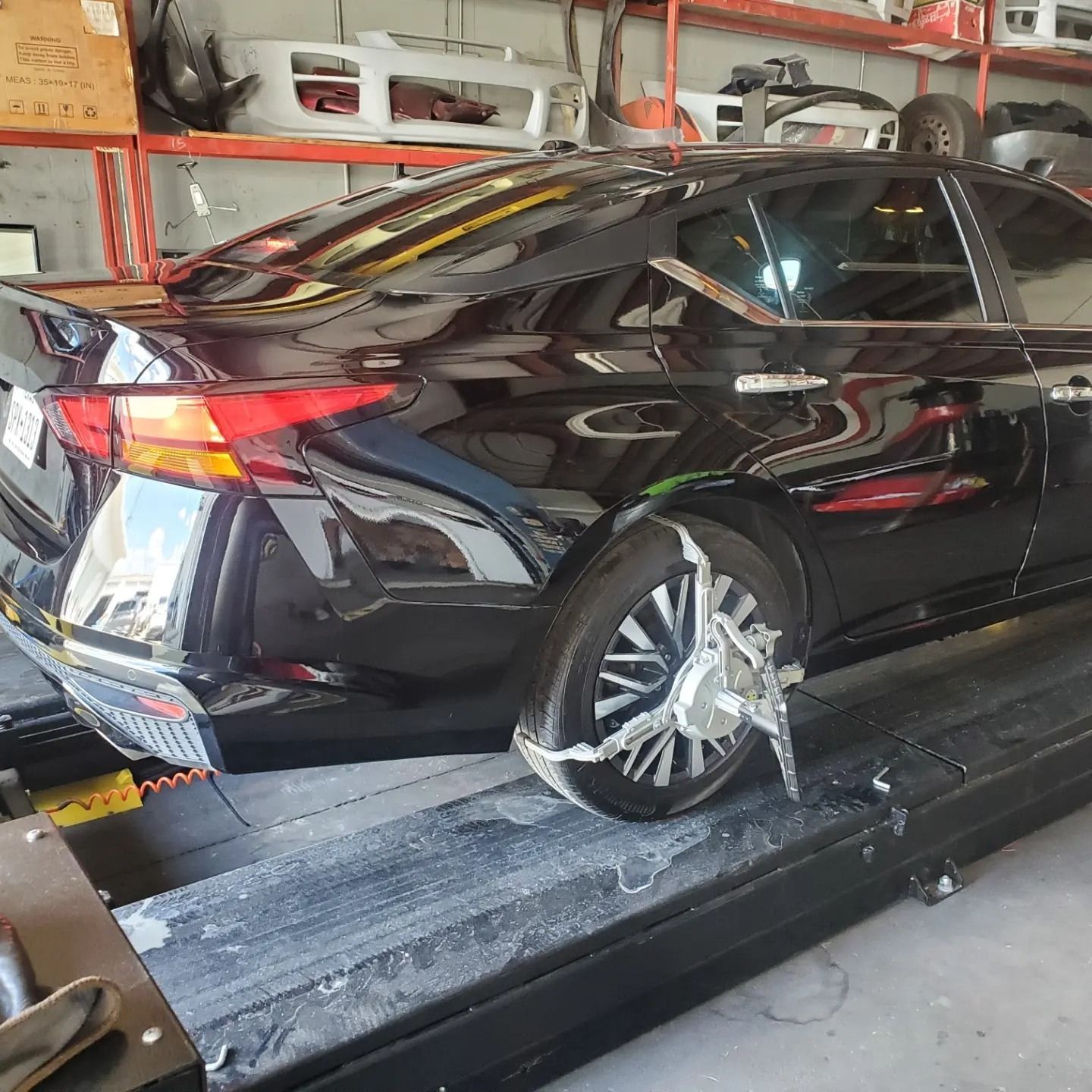 Black sedan on a dynamometer in an auto shop, with a damaged rear wheel and body damage visible