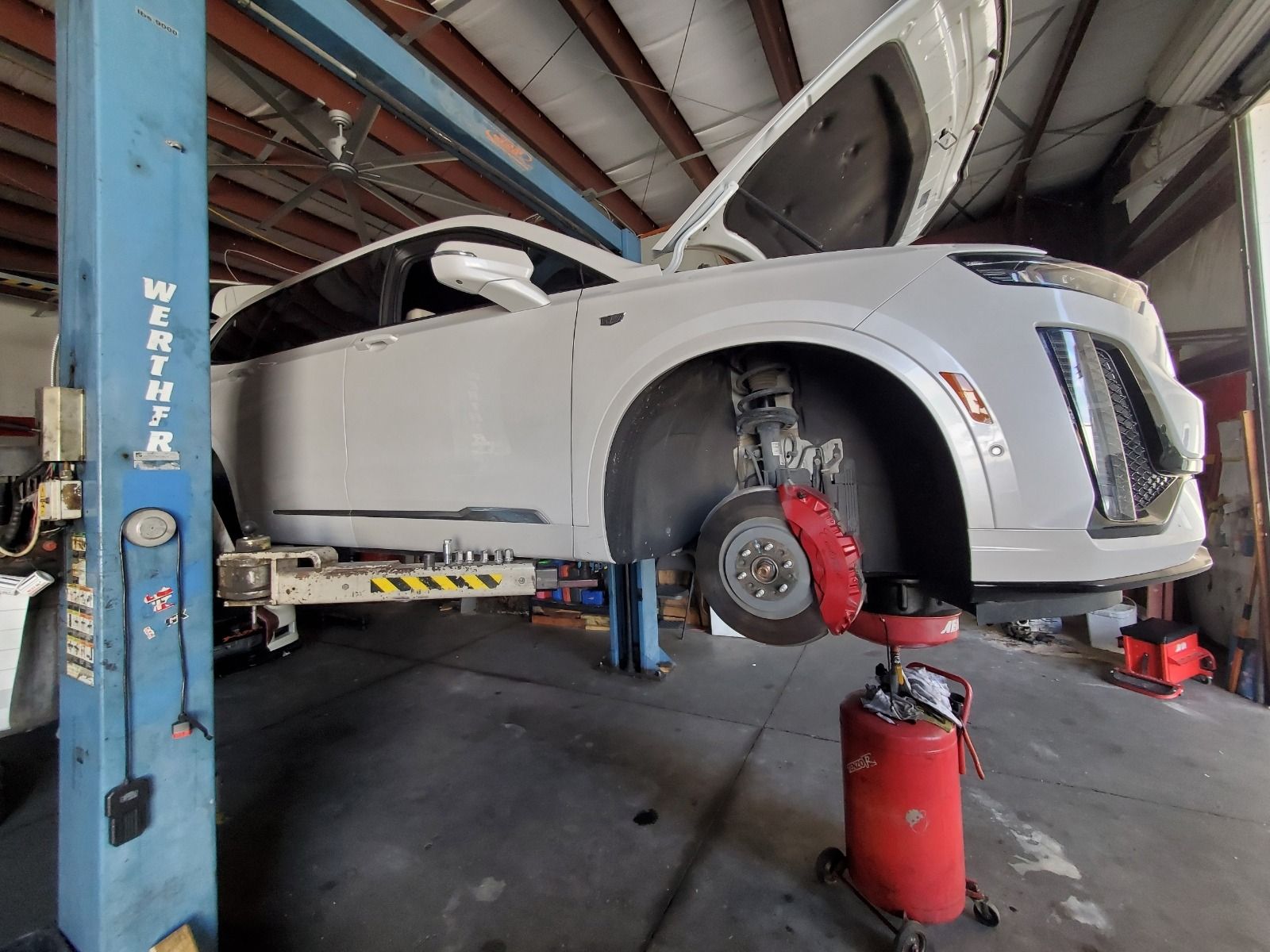 White SUV lifted in a repair shop, front wheel removed with brake exposed on a red jack.