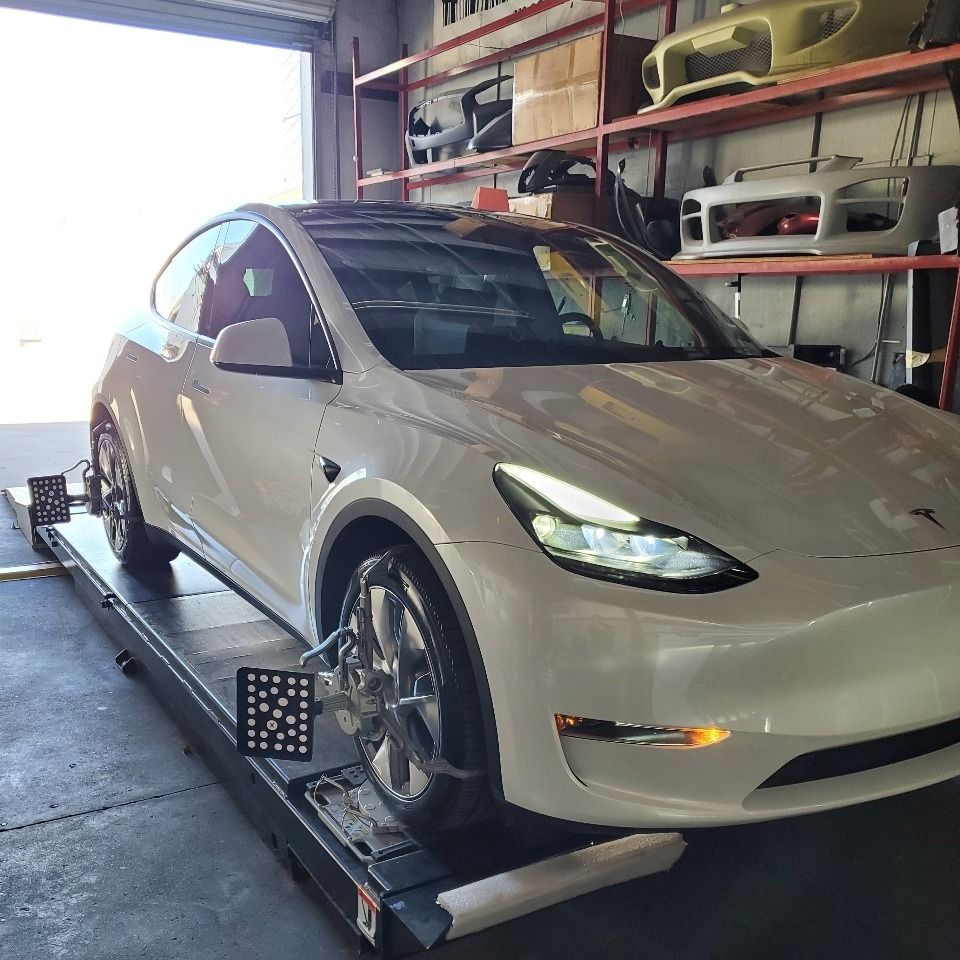 White Tesla on a vehicle lift inside a garage, with front lights on.
