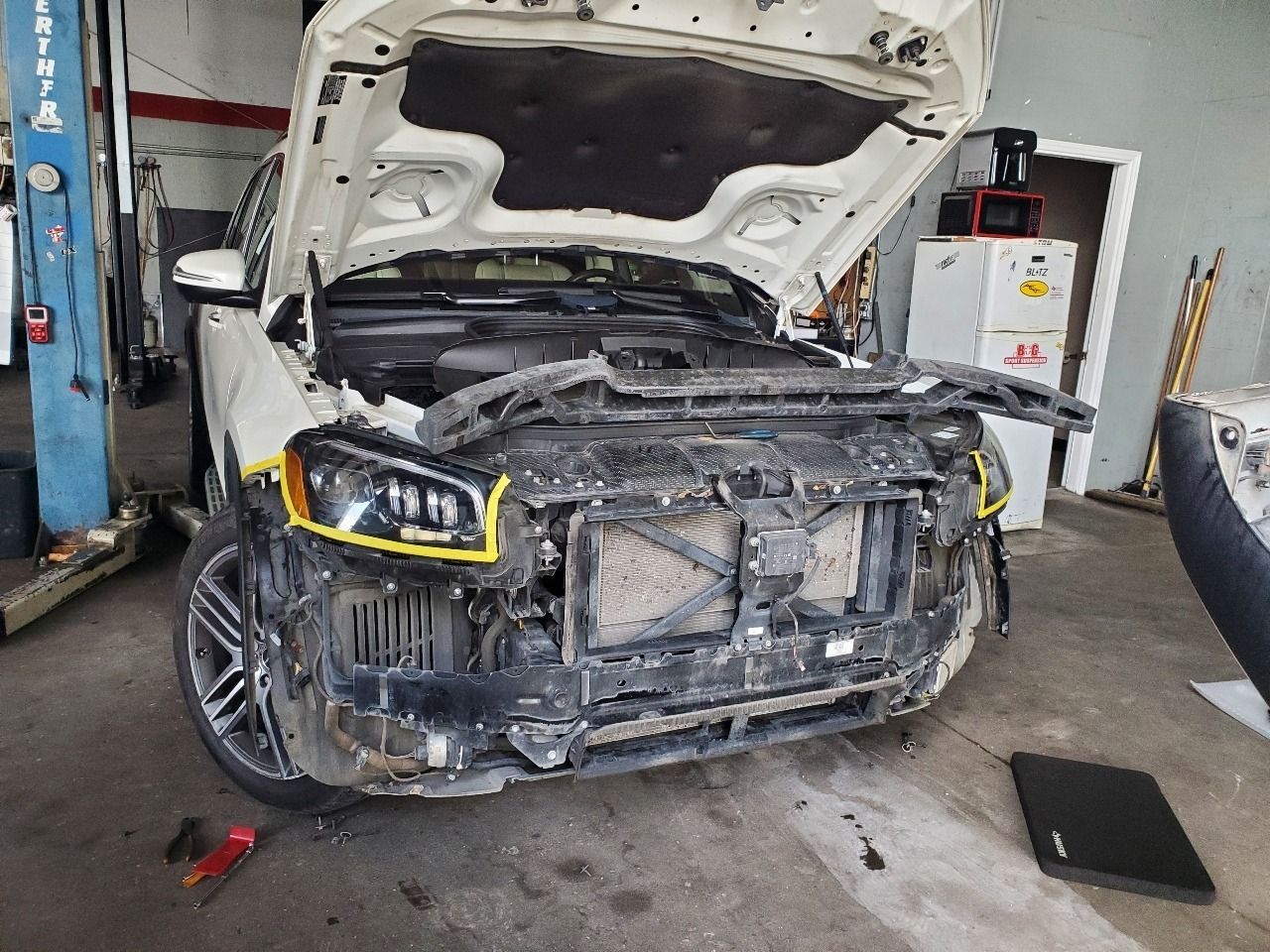 Damaged black car with hood open in an auto repair shop, front bumper removed and headlight highlighted.