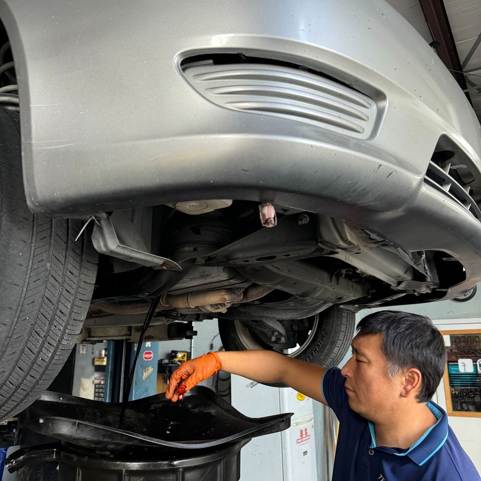 Mechanic in orange gloves draining oil from a car on a lift in an auto repair shop