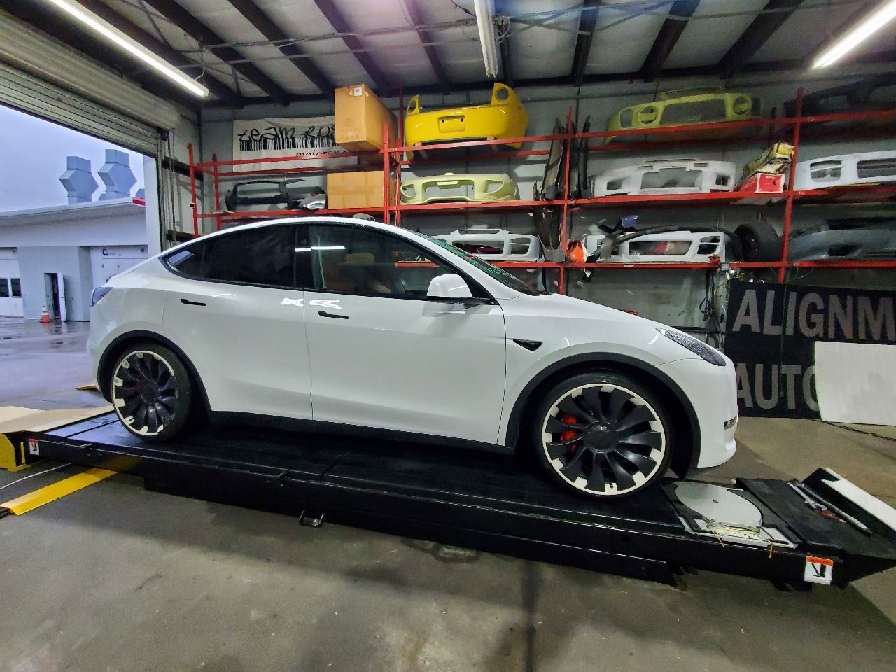 White SUV on a car alignment lift inside an auto shop with tires and wheels on wall racks