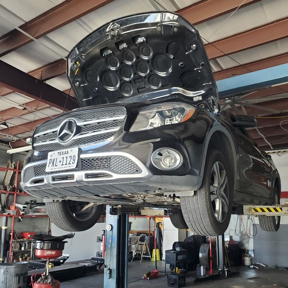 Black Mercedes SUV raised on a car lift in an auto repair shop, hood open and front wheels hanging.
