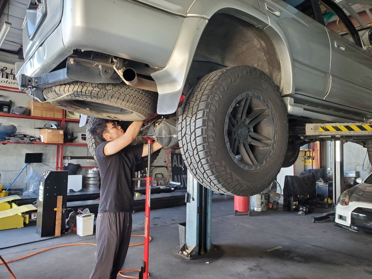 Mechanic working under a lifted silver SUV in an auto shop, checking the exhaust.