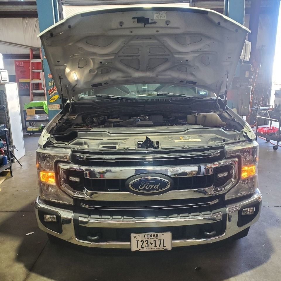 Front view of a silver Ford truck with its hood open in a garage, headlights on.