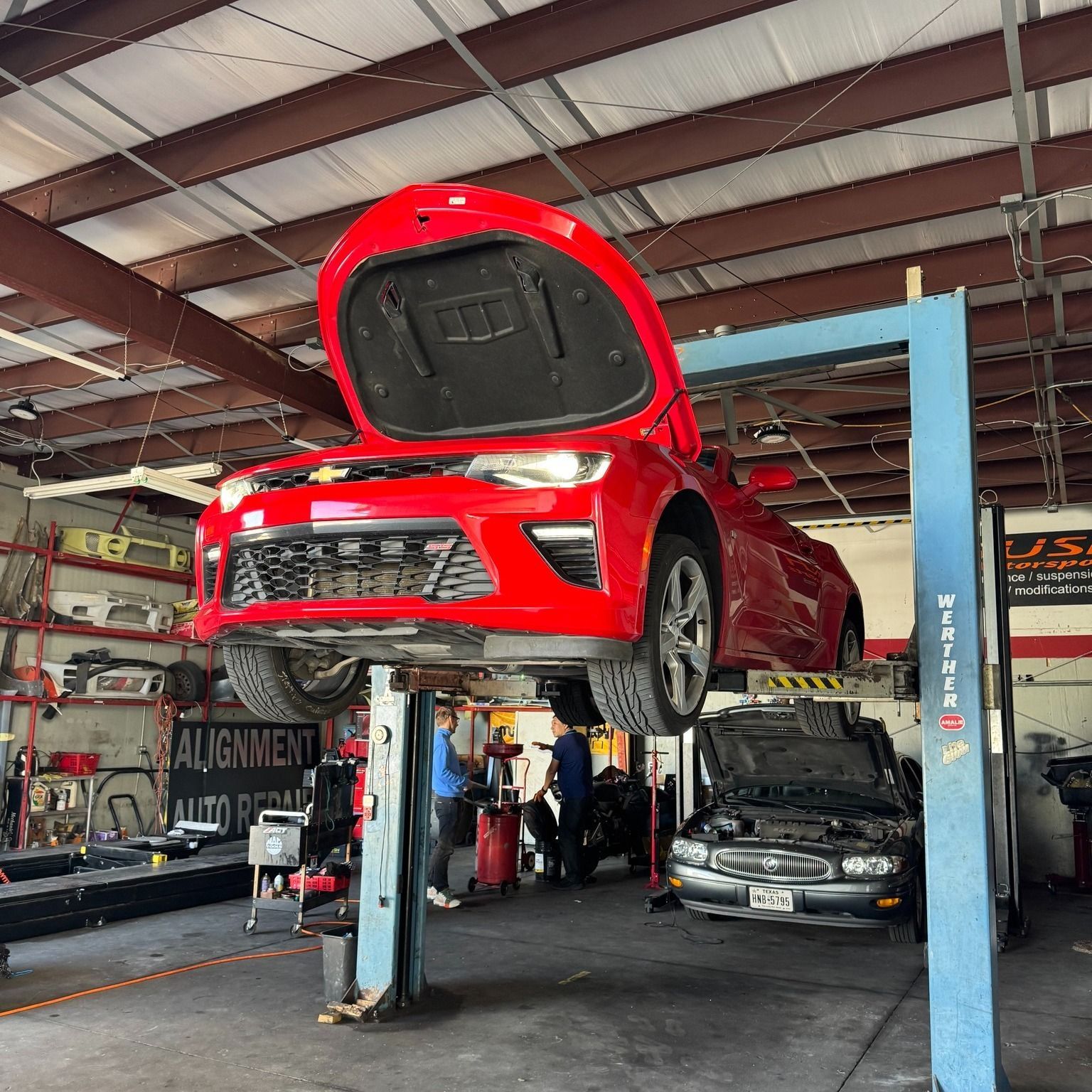 Red car lifted in auto shop with hood open, mechanic working nearby, another car under repair beneath it