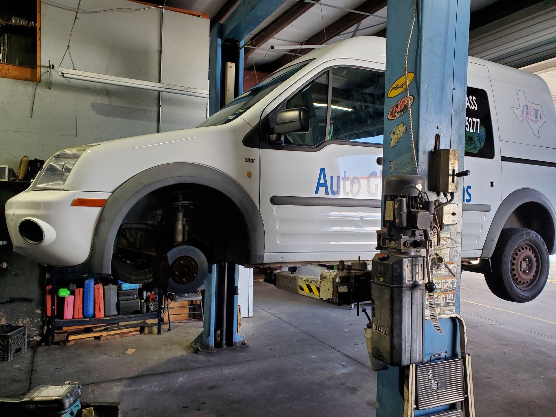 White van lifted on a car jack in an auto repair shop, with front wheel removed.