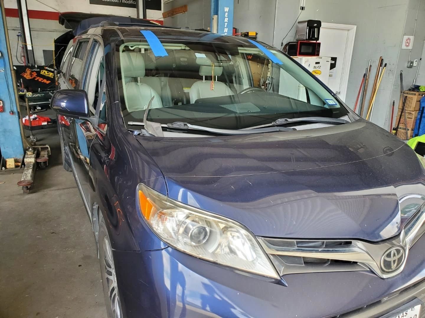 Blue Toyota minivan parked in a garage workshop, front view with windshield tape.