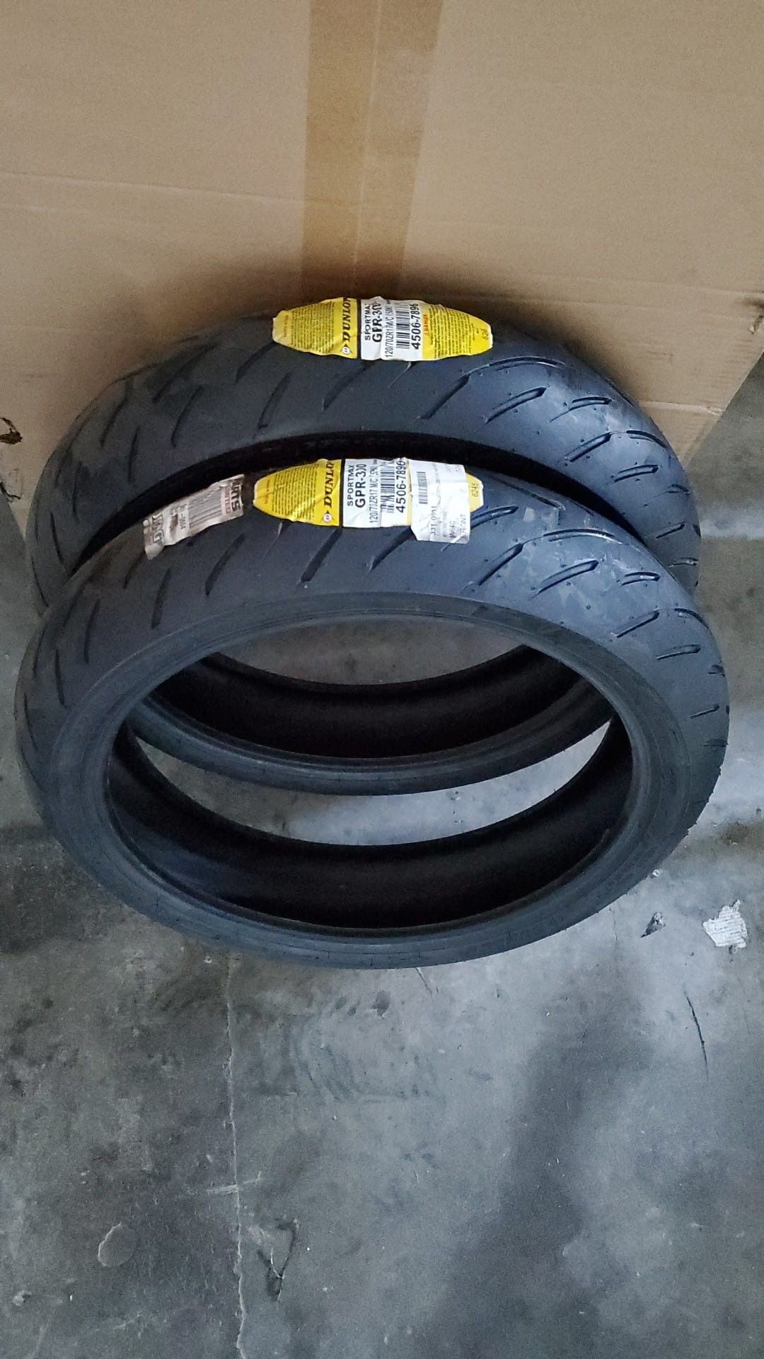 Two stacked black motorcycle tires with yellow labels on a cardboard box and concrete floor