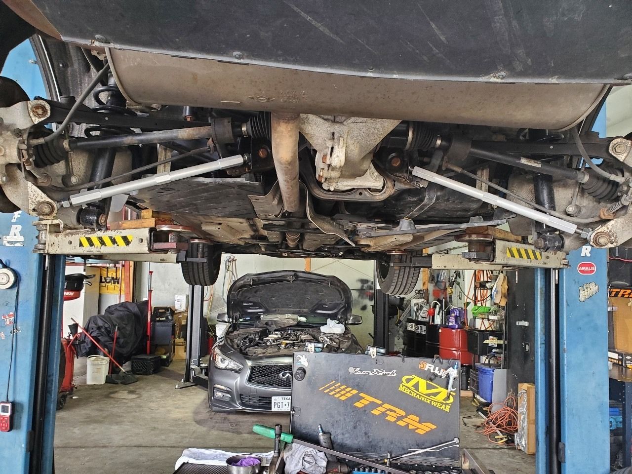 Car on a lift in an auto shop, underside exposed, with another car and tools visible below.