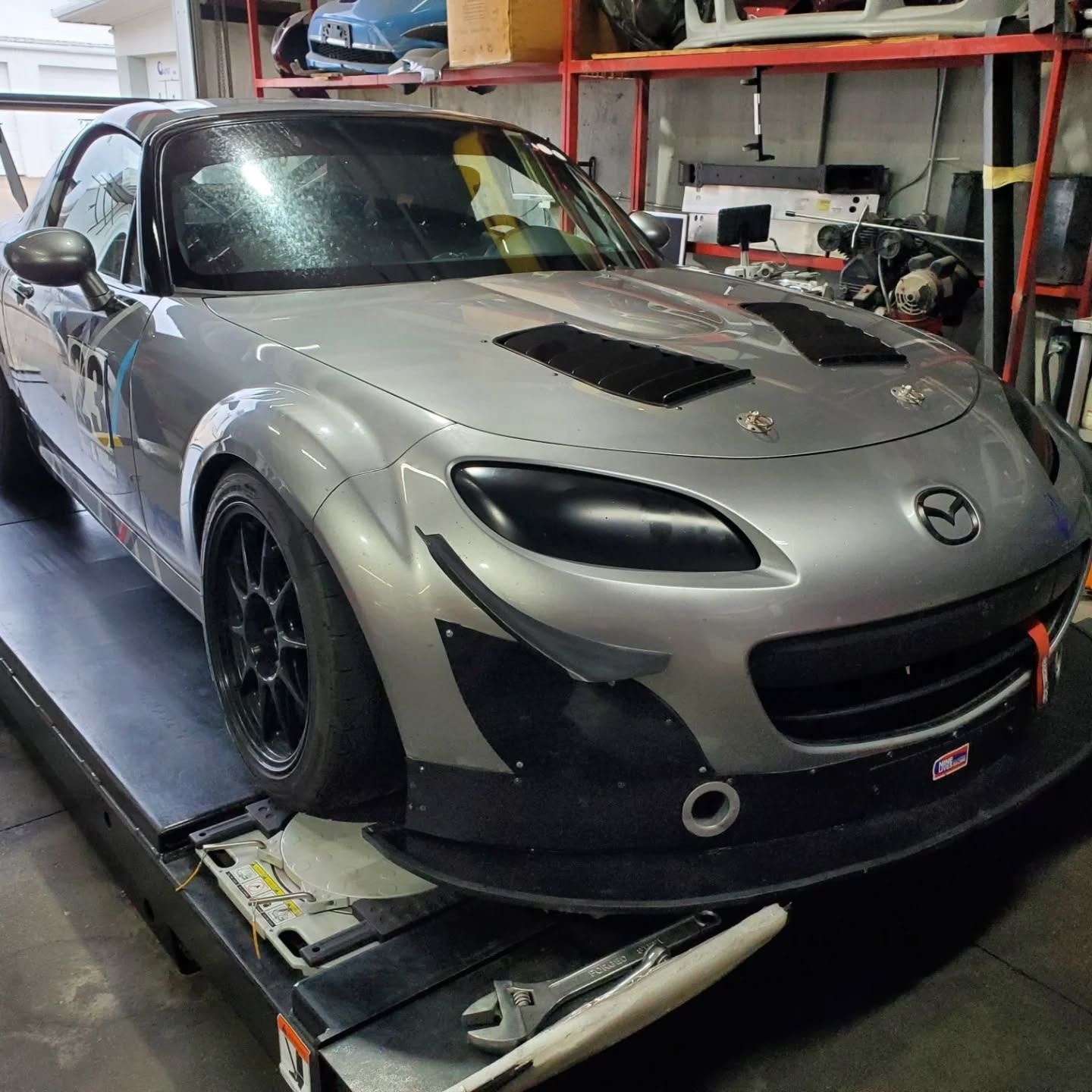 Silver Mazda race car on a shop lift in a garage, with black hood vents and front splitter