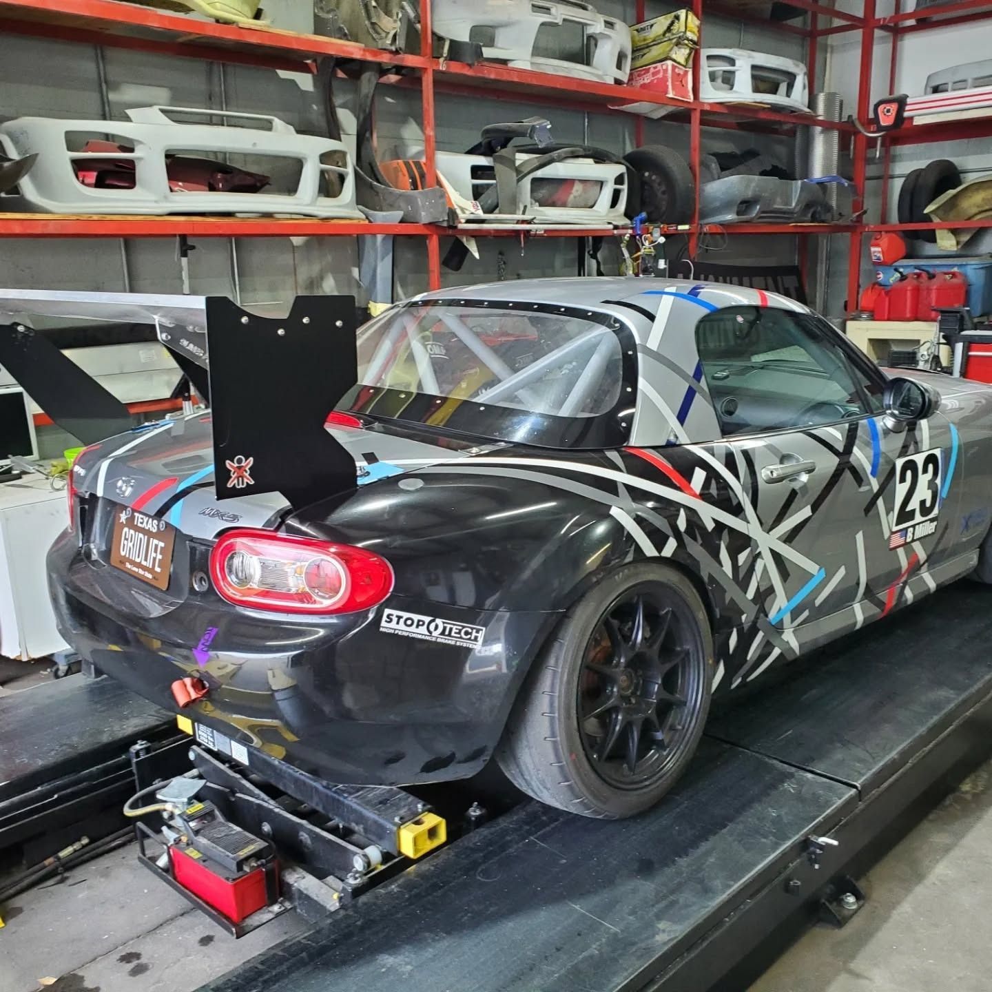 Race car with black-and-gray camo wrap on a lift in a garage, viewed from the rear.