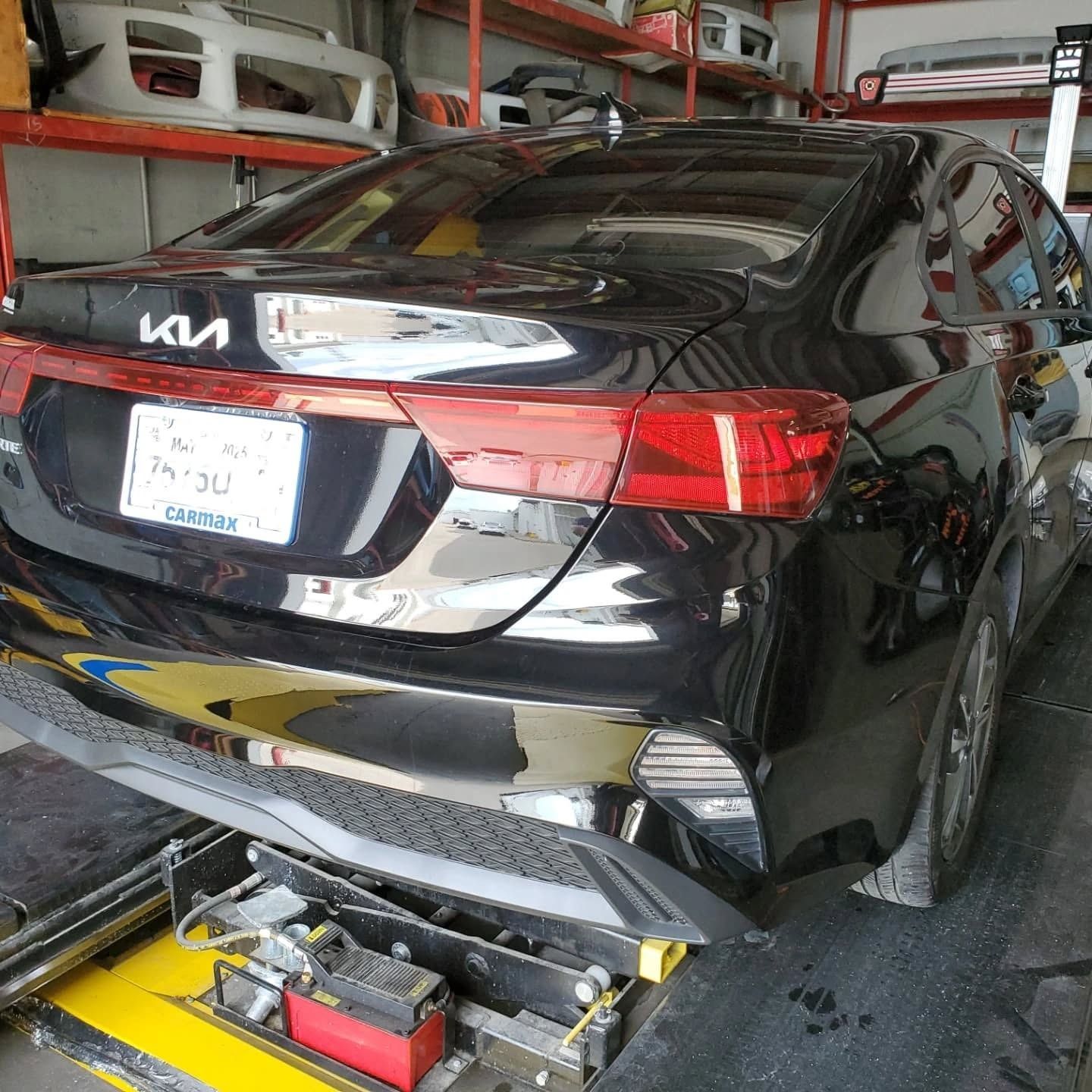 Black Kia sedan on a car lift in a repair garage, viewed from the rear.