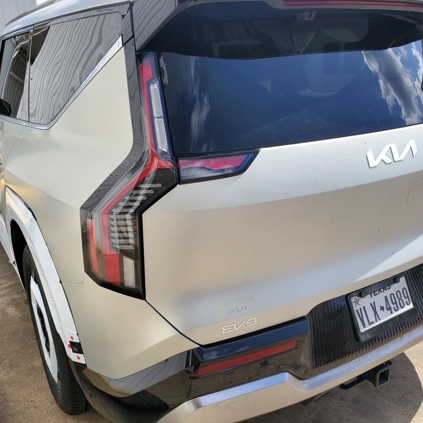 Rear view of a beige Kia SUV with damaged left taillight and dented bumper in a parking lot