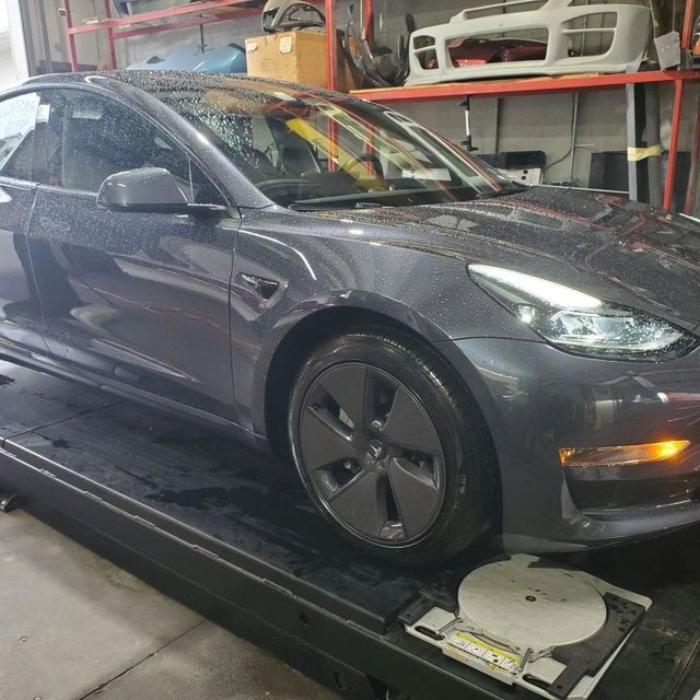Dark gray Tesla sedan on a lift in a garage, with white body parts on shelves behind it.