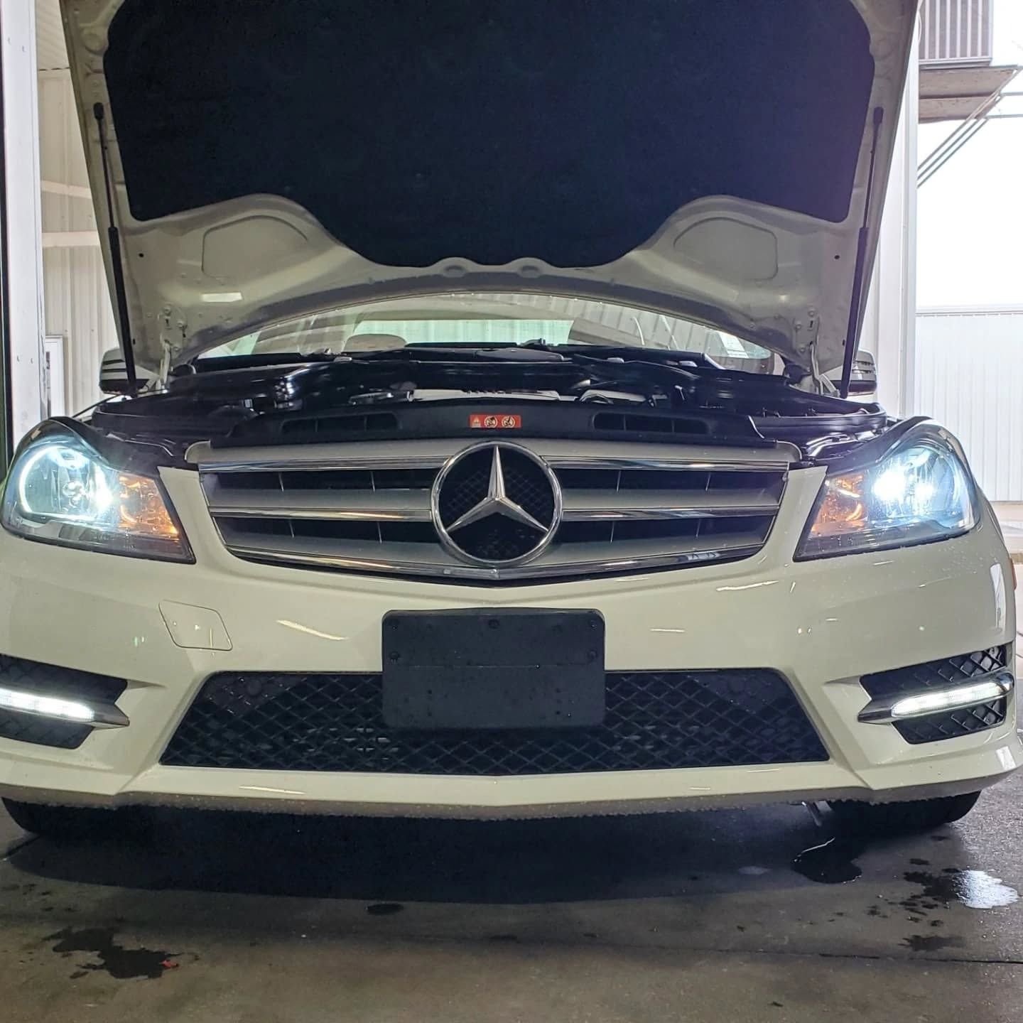 Front view of a white Mercedes-Benz with hood open and headlights on in a garage.