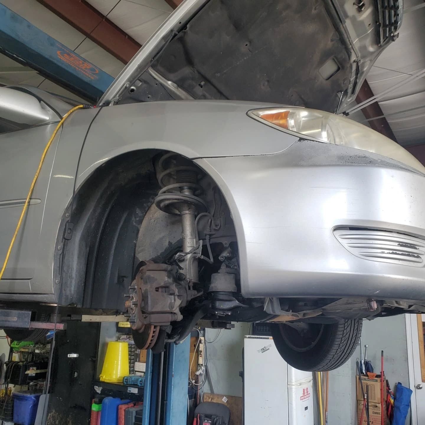 Silver car raised on a lift in an auto repair shop, showing the front wheel removed and exposed suspension.