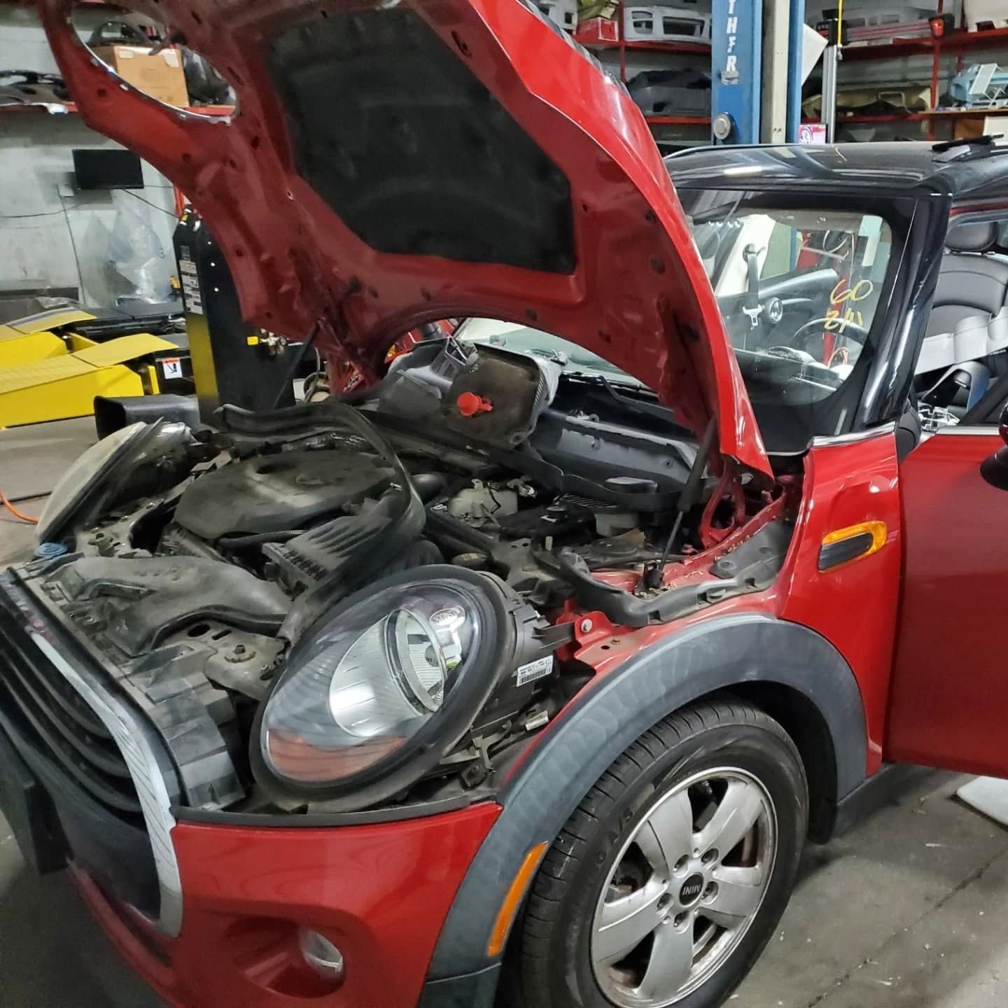 Red Mini Cooper with hood open in a repair shop, showing exposed engine parts.