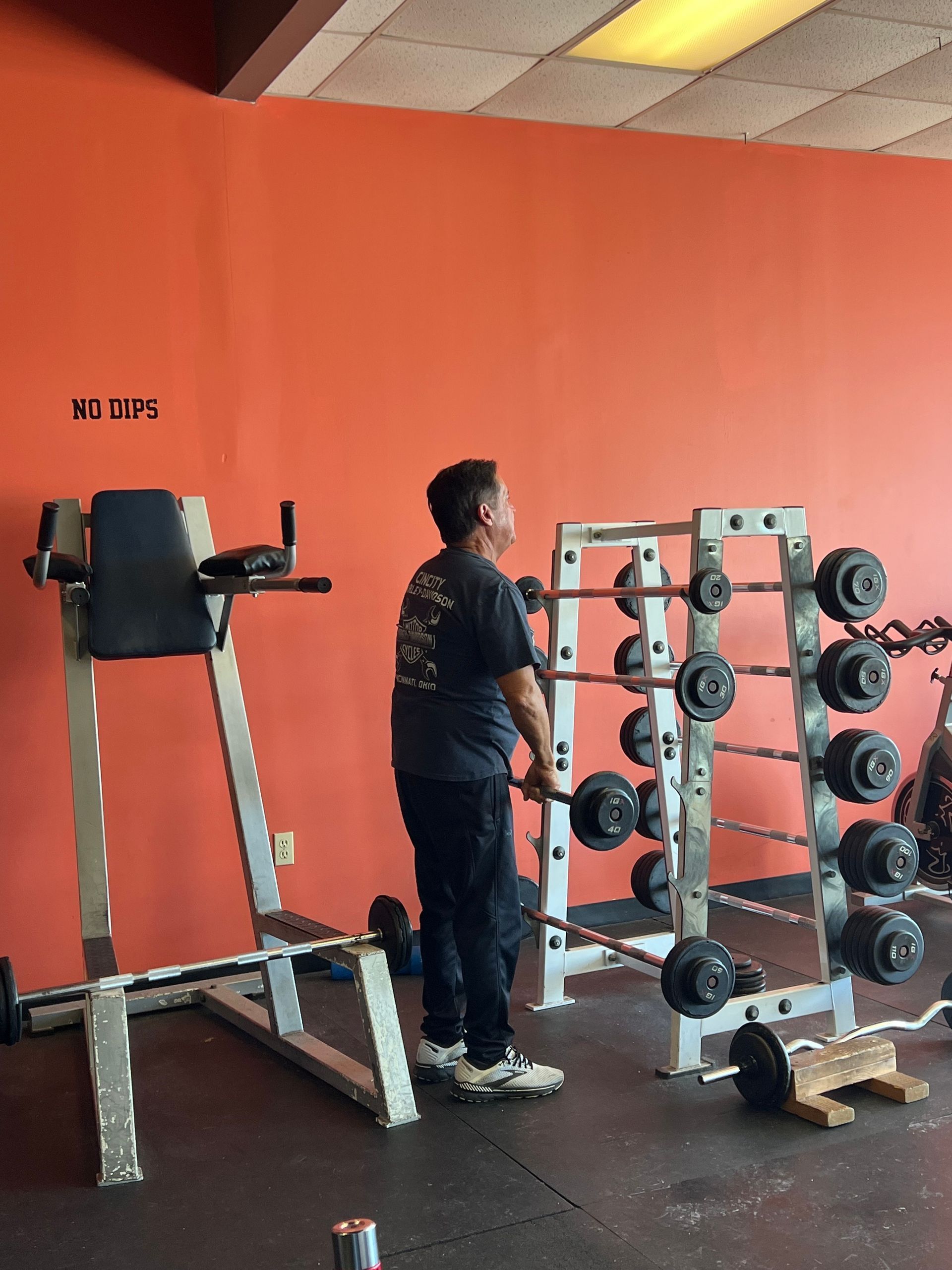 Person in a gym, looking up at weight rack. Orange wall in the background. Weight lifting equipment visible.