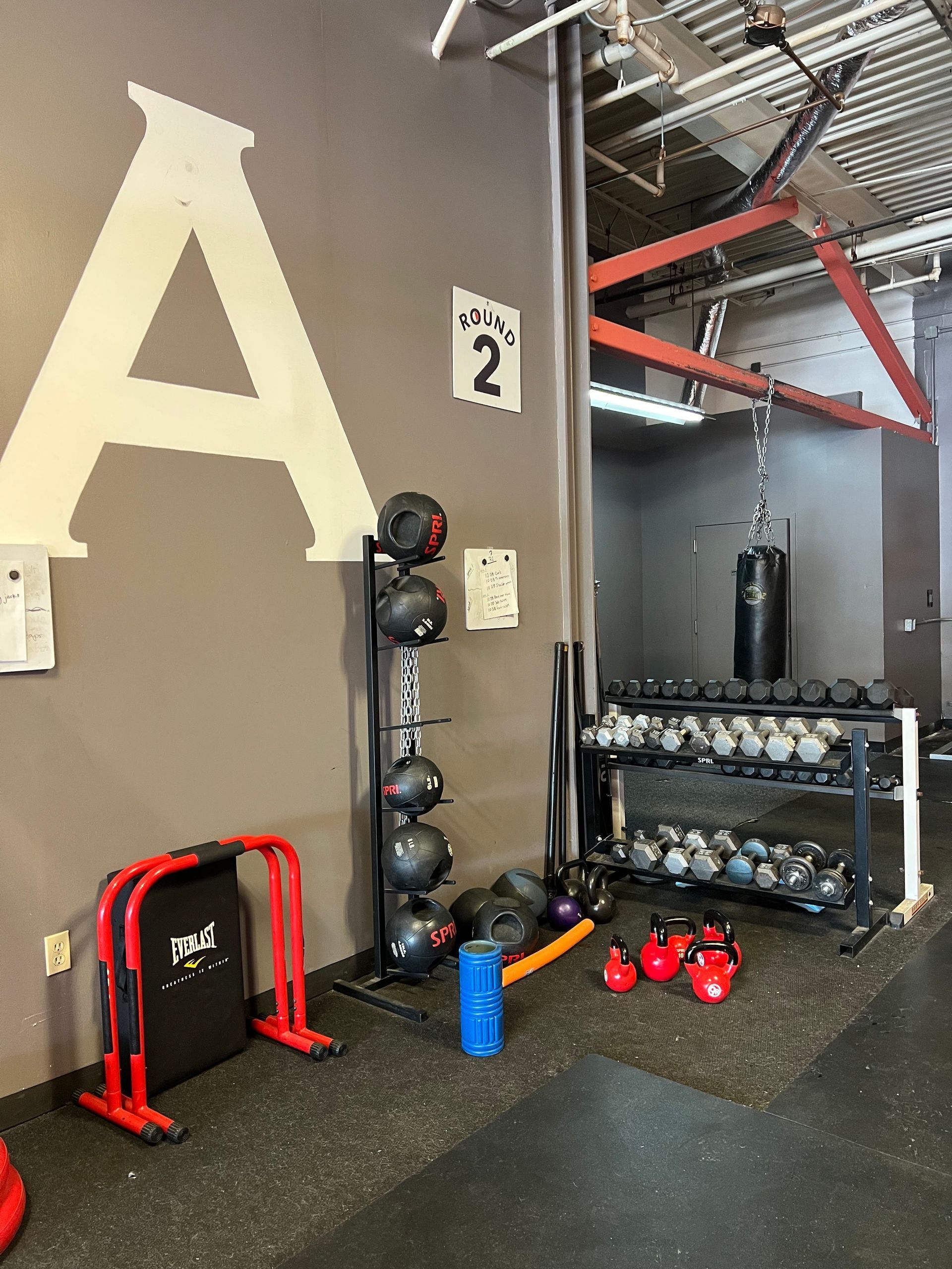 Gym interior: weights, kettlebells, punching bag, and exercise equipment. Gray wall with a big letter A.
