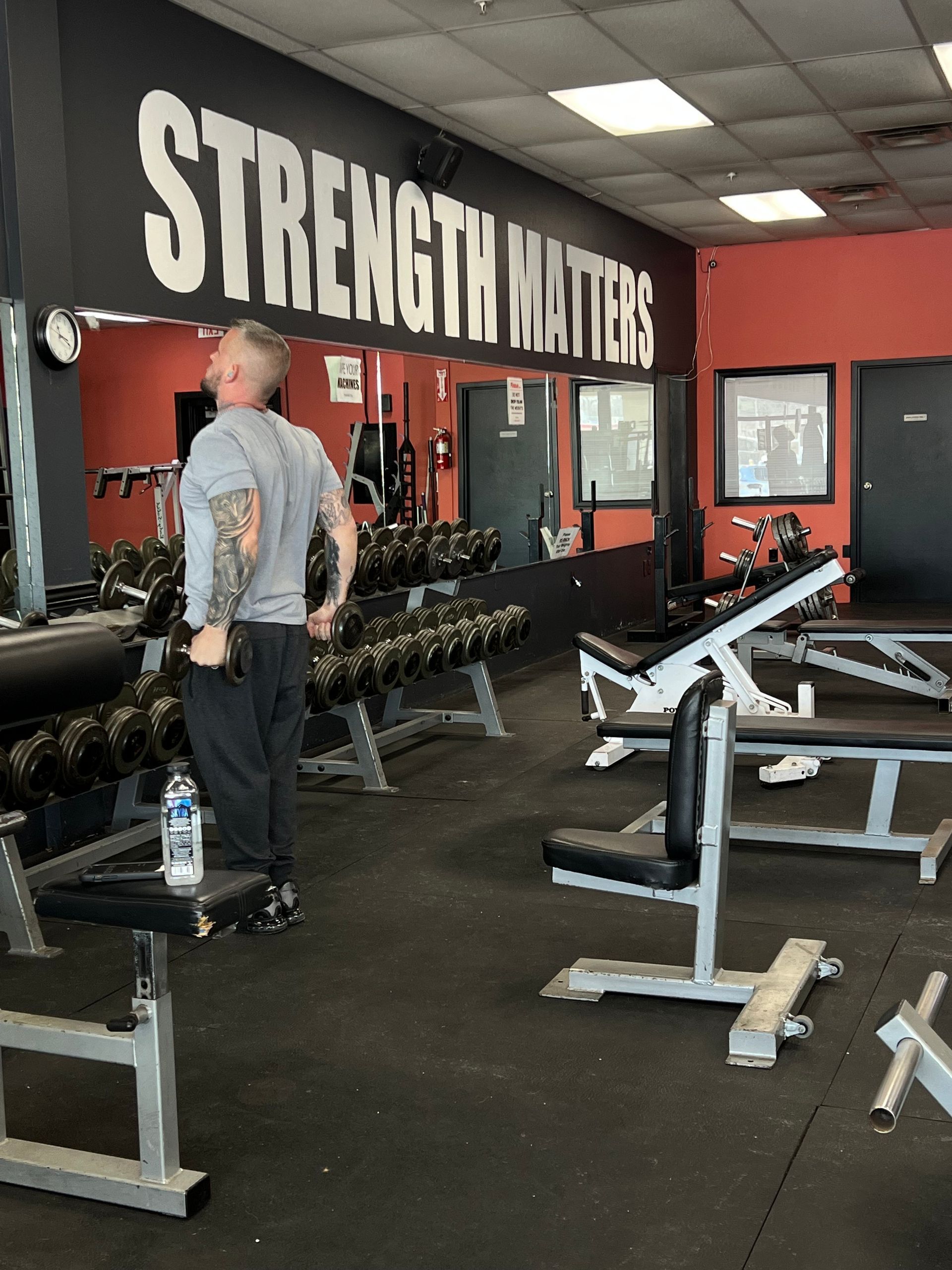 Man in a gym stretches, surrounded by weights and benches. Wall with text: Strength Matters.