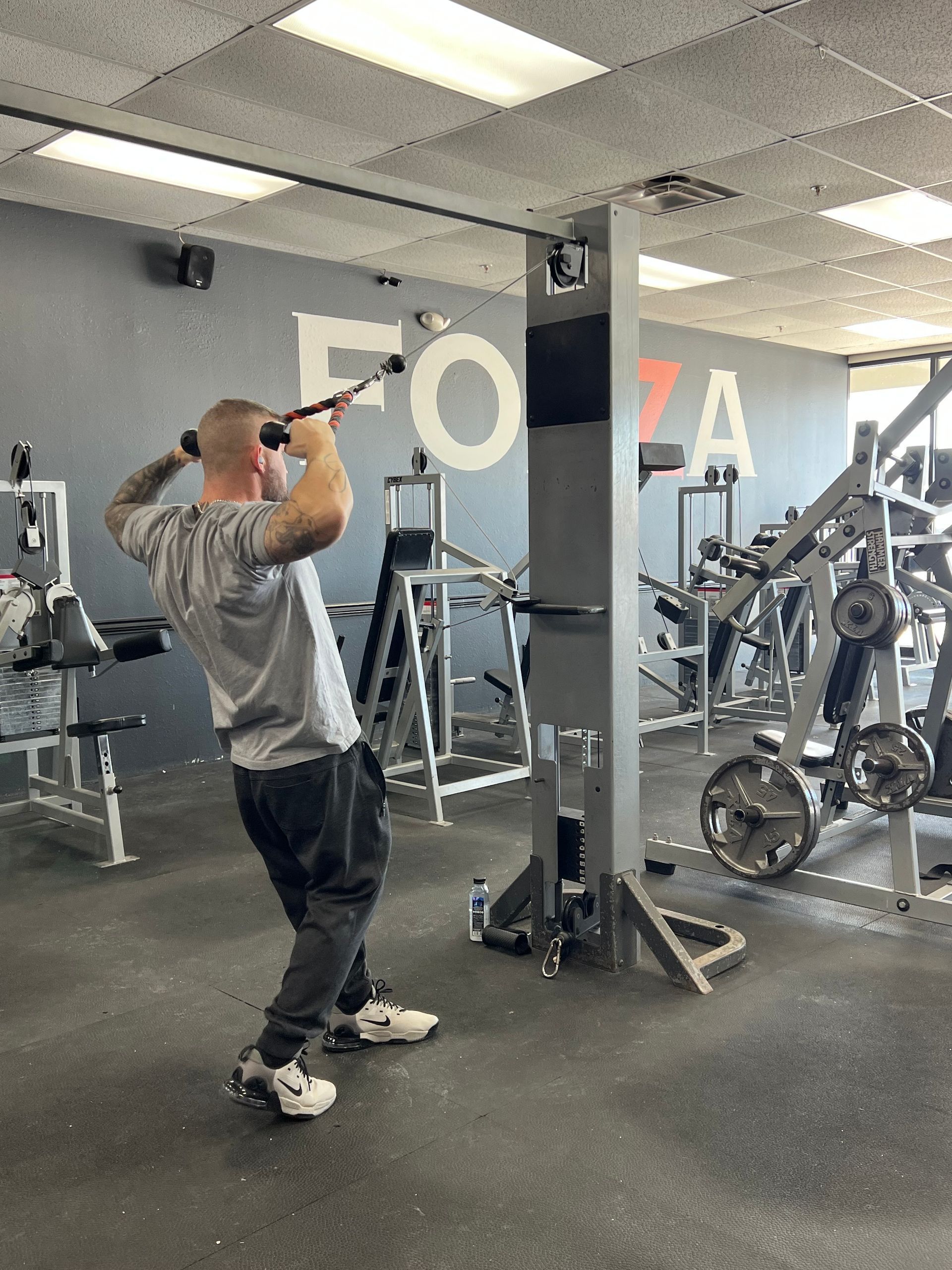 Man in gym doing cable triceps extension. Grey shirt, dark sweatpants, white sneakers. Gray and black gym equipment.