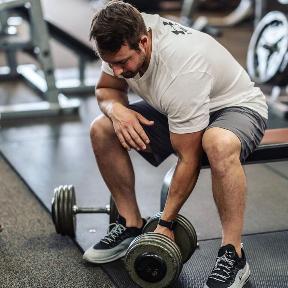 Man in gym shorts and t-shirt rests arm on a dumbbell in a gym.